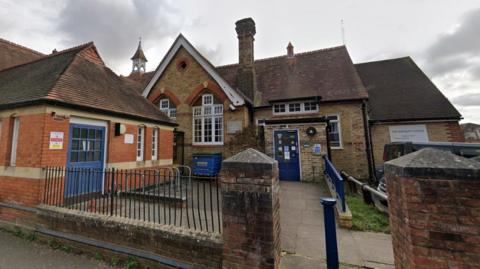 The Maybury Centre Trust building, made up of red and brown bricks with blue blues, stands below a grey and cloudy sky. A brick fence with black metal bars sits in front of the building.