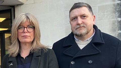 Liz Hopkin (left) on the right with blonde hair and glasses, standing outside Swansea Crown Court with her husband John, on the right. He has got short grey hair, a beard, and is wearing a dark navy coat.