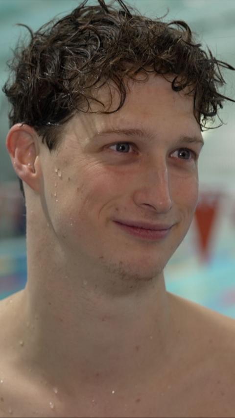 A swimmer with damp, short brown hair smiles as he looks to the side of the camera. He is standing poolside.