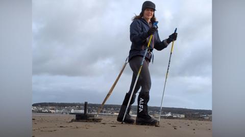 A woman with long brown hair and hiking boots is dragging a sled along a beach with the Grand Pier in Weston-super-Mare in the background. The sled is connected to her waist. She is holding walking poles.