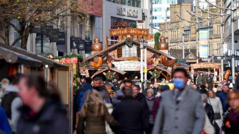 several shoppers walk past wooden chalet-style stalls decorated in traditional wreaths and items on Cathedral Street, surrounded by shops