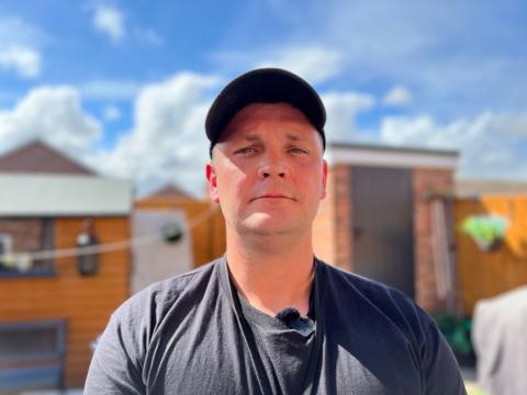 Ashley Waines is wearing a black t-shirt andbalck baseball cap and is standing in a garden with a wooden shed visible behind him