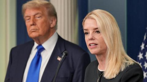 US Attorney General Pam Bondi, a woman with short blonde hair wearing a black blouse, speaks in front of a US flag, with US President Donald Trump behind  her wearing a dark blue jacket, white shirt and bright blue tie.