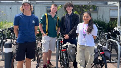 Four students standing in front of a bike rack. There are three boys and a girl, they are all looking at the camera and smiling. 