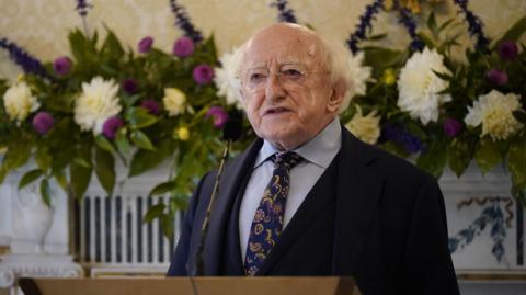 Michael D Higgins wears a navy suit with a blue shirt and purple and navy tie. He stands at a brown plinth with a black mic attached to it. Behind him on the cream wall is a green white and purple flower arrangement that stretches horizontally across the whole frame.