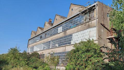 An exterior view of the derelict Zig Zag building in Glastonbury. It appears to be about three storeys and has five separate pointed triangular roof structures. It is built of grey concrete with lots of square glass windows on each floor.