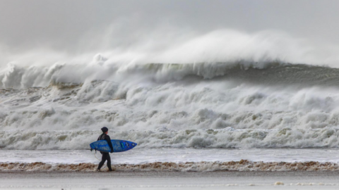 A person holds a surf board whilst waves are high nearby.