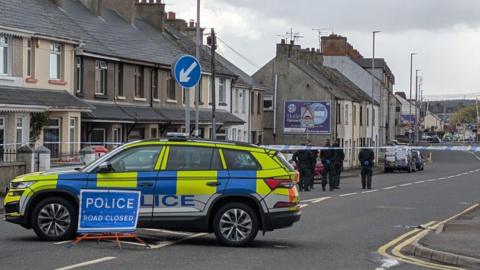 Yellow blue and white police vehicle at the scene of a shooting on the Bushmills Road in Coleraine. There are police officers dressed in black in the distance and houses to the left.