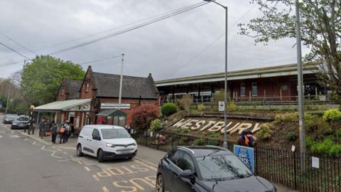 The front of Westbury railway station, showing Westbury in large letters in a flower bed and a traditional red brick building, with the tracks raised up behind. A few cars in a pick-up area at the front