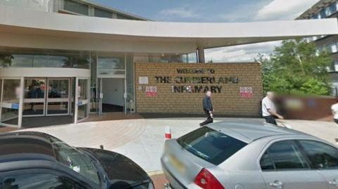 The entrance of Cumberland Infirmary. It has white revolving doors and a sign reading Welcome to Cumberland Infirmary. Two people are walking past and there are two parked cars.