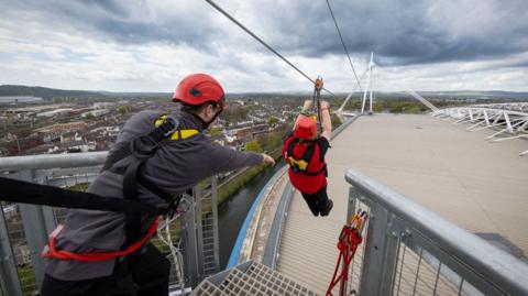 The top of the principality stadium from a platform used to launch the zip liner. A man on the left of the photo is strapped on with a black strap. His arm is outstretched towards a woman who has just launched. He is hanging on to a handle next to the zip line and roller. She is strapped in with a shoulder harness visible. The roof of the stadium and the city beyond are in the background.