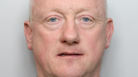 Mugshot of Christopher, against a grey backdrop. He has short white hair, blue eyes and thin eyebrows.