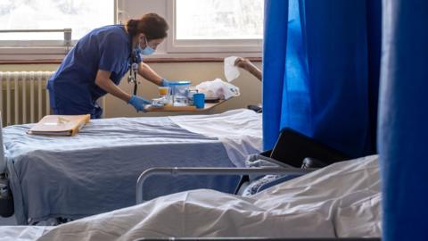A hospital ward with several beds separated by blue privacy curtains. A healthcare worker in blue scrubs and a face mask leans over a bedside table while attending to a patient beside a bed. Large windows provide daylight, and medical equipment and bedding are visible throughout the room.