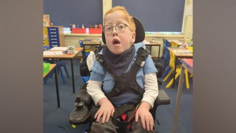 Ezra, sat in an electric wheelchair and looking at the camera. He is within a colourful classroom with small chairs and desks.