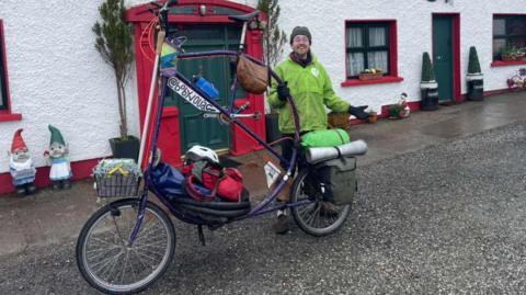A man is standing beside a large bicycle. The seat and handlebars are taller than his head. The bicycle is painted purple and has baskets and bags attached on the front and back. The man is wearing a green coat and hat. 