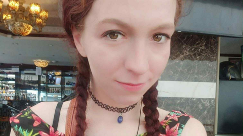 A selfie of a young woman with long red hair in two plaits. She is sitting smiling in front of a bar. 