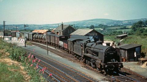 An archive image of a steam train with more than 14 freight carriages, being loaded at Bere Alston Train Station. 