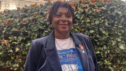 Beverly Douglas. She has short dark hair and is wearing a navy blue blazer with a poppy badge on the collar. She is also wearing a white t-shirt with a blue box that reads her name and PC Bev. She is looking directly at the camera and smiling.