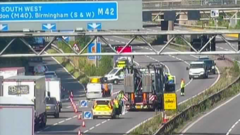 Police close off the motorway, with vehicles, signs and cones blocking the carriageway and directing a long stream of traffic on to a slip road