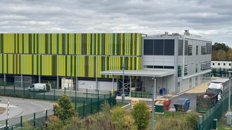 A data centre that looks like a large warehouse surrounded by tall, dark green metal fences. The building looks like it is largely covered in green stripes which run vertically down from the flat roof. The stripes are fins which help the cooling of the centre. They are in various shades of green. On the right of the warehouse are some windows which serve a number of offices inside.