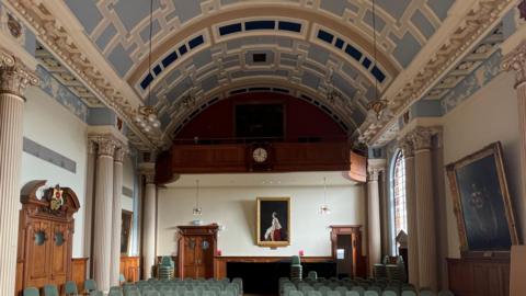 Ornate geometric pale blue and cream curved ceiling in room with pale green seats. Wooden panelling and a clock can be seen on one wall and ornate a wooden doors. A portrait of the late Queen Elizabeth hangs on one all with other artworks. 