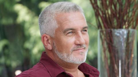 Paul Hollywood is seen in a closeup photograph on a baking show. He is wearing a shirt and has grey hair and a beard and he is smiling. There is a vase of stems behind him from a flower arrangement.
