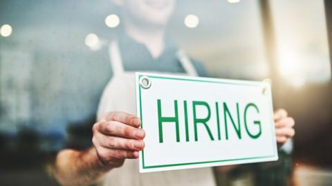 A man holds a white sign with green writing which says "Hiring"