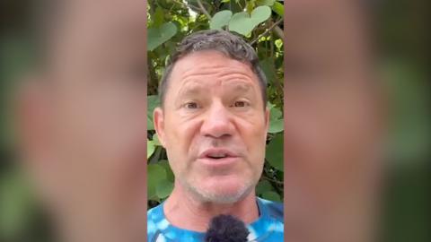 Steve Backshall stands in front of some foliage. He is speaking into a mic and wearing a mottled blue T-shirt.