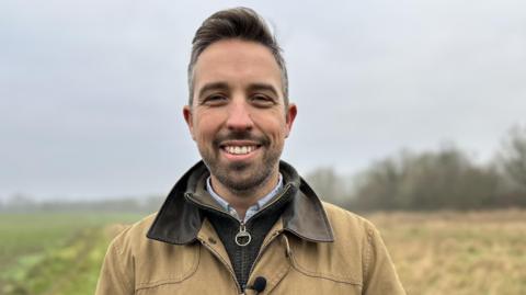 Tristan Baxter-Smith in a tan coloured jacket stands in a field smiling. He has greying hair and stubble.