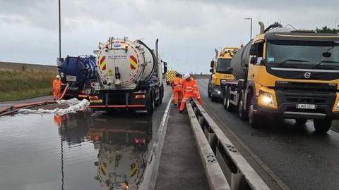 Road with central reservation - to the right are several yellow lorries and on the left are tankers with workers in orange suits and brown water is gushing into the road