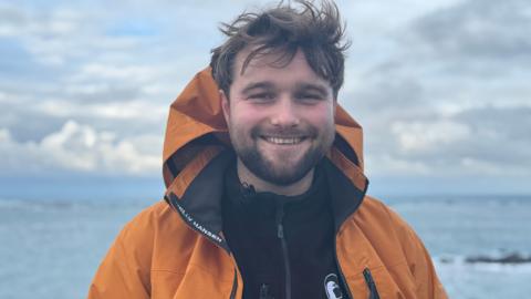 Alex Purdie standing in front of the coast, smiling. He has brown hair and a beard. He is wearing an orange coat over a zip-up fleece with the Alderney Wildlife Trust logo on it.
