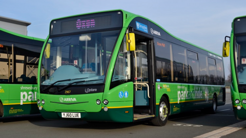 A number of green park and ride buses. The central one has the word Arriva on the front and its door is open and no-one is sat behind the wheel of the stationary vehicle