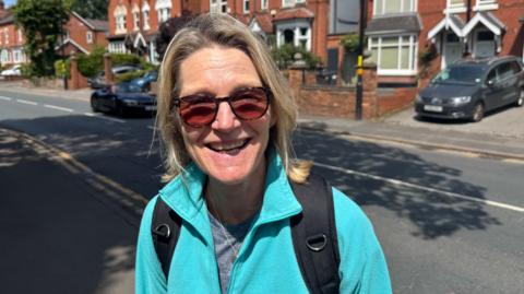 Dee Lyons in a smiling photo. There is a road behind her with a car driving past. She is wearing a light blue fleece and sunglasses. She has blonde hair. 