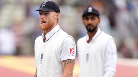 Ben Stokes, wearing a blue cap and white sweater, looks frustrated, left, with Shoaib Bashir in the background