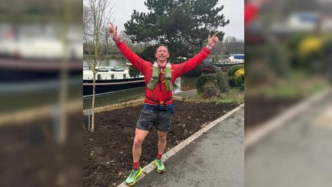 A middle-aged man wears a red training top, dark grey shorts, red socks and green trainers, as he stands in a park with both hands raised in triumph above his head.
