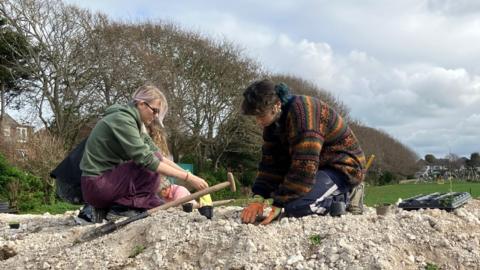 Two women planting a wildflower bank at a park. They have shovels and other gardening equipment with them.