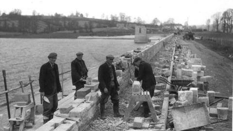 A black and white photo of four men working on some brick work by the edge of the water. They have flat-caps on and are dressed smartly in wellies and dark clothing. You can see the wall of the edge of Chew Valley gradually being built with the water tower in the background.