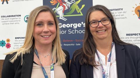 Two women stood side by side, smiling and looking into the camera. Kelly, on the right, is wearing a black blazer and a beige top. Sarah, on the left, is wearing a black blazer and a white shirt. They are both wearing blue school lanyards and are stood in front of a white sign with words like 'collaboration' 'empowerment' 'positivity' and 'respect' written on it in blue.