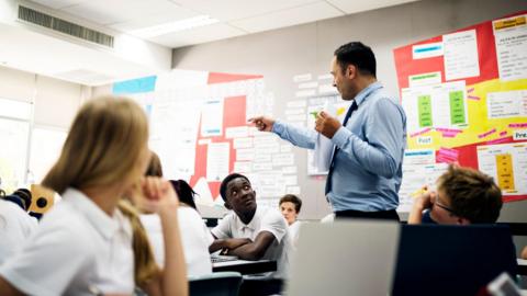 A stock image shows a secondary teacher standing teaching in a classroom wearing a suit and tie. The students are looking at him from their desks, wearing white short sleeved shirts.