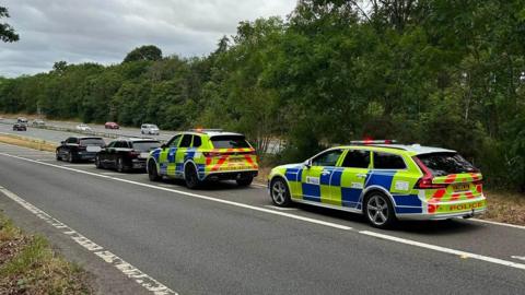 Police vehicles with flashing lights are parked on the roadside of the A46 near a wooded area while officers carry out a traffic enforcement operation, with several stopped cars lined up ahead under an overcast sky.