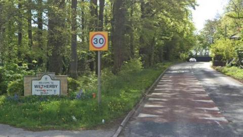 A road stretches ahead with trees and grass verges on either side. On the left is a 30mph road sign and a sign saying Welcome to Wetherby on a low stone wall.