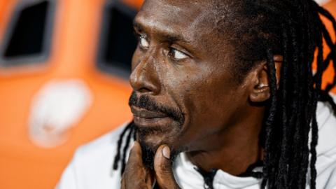 Aliou Cisse, with his back hair braided and a goatee beard, holds his left hand to his chin as he looks to his right