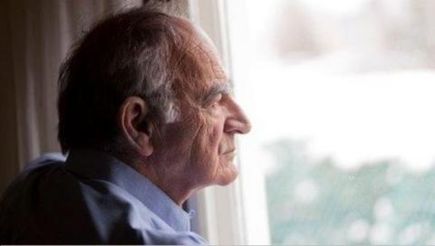 An elderly man wearing a blue shirt, looking our a window