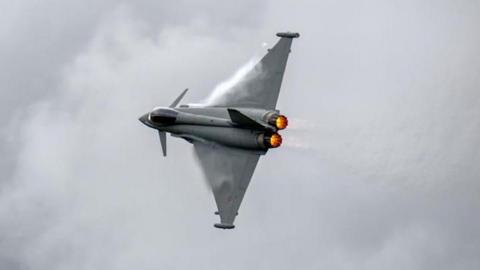 A grey aircraft with long wings and fire at its tail in front of a grey sky covered by white clouds.