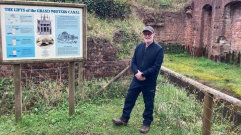 A man dressed in navy walking gear, walking boots and a baseball cap stands in front of a heritage site on the former Great Western Canal route