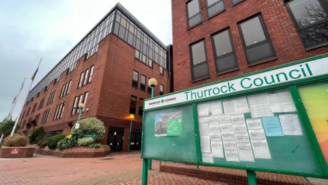 A large green sign and notice board that reads "Thurrock Council" at the top. It is placed in front of two brown-bricked multi-storey buildings. 