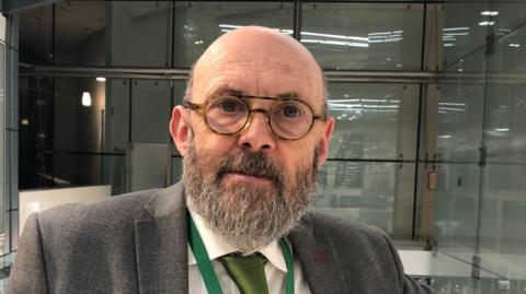 Andrew Stringer looks at the camera. He is bald with a long dark grey beard and glasses. He wears a grey suit with a green tie and a green lanyard around his neck.