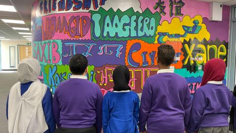 Students at the school with their backs to the camera, looking at a graffiti wall with greetings in different languages. They are wearign purble and blue jumpers.