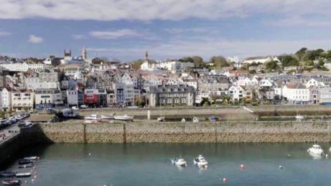 Guernsey harbour with water in the forefront and a number of houses in the back, with blue sky at the top 