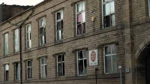 A two-storey stone-built commercial building with multiple broken and damaged windows is shown from street level. Numerous window panes on both floors are shattered or missing entirely, with some openings partially covered by curtains, boards, or remaining fragments of glass. Electrical cables are visible running across the exterior wall near the upper floor. A weathered sign mounted on the front of the building reads "new weighing line" with a telephone number beneath it, although parts of the text are difficult to read. The building appears vacant or neglected, and the surrounding stonework shows signs of age and wear. An arched stone entrance is partially visible on the right-hand side of the image.